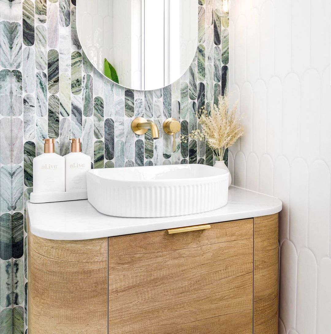 Modern bathroom vanity setup featuring a wooden cabinet with a curved design and gold handle, a white ribbed countertop basin, and wall-mounted gold taps. The backdrop showcases an elegant tiled backsplash with green, white, and earthy tones in an elongated oval pattern.