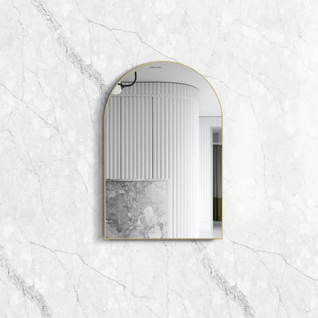 Archie 900x600 Gold Metal Framed Mirror with an elegant arched design, reflecting a modern interior against a polished white marble-textured background.