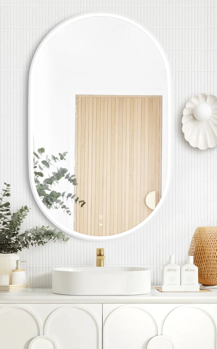 LED Noosa Mirror in 900x600 dimensions with a white metal frame, mounted above a modern white vanity, accented by a round vessel sink, gold faucet, and natural décor elements including eucalyptus and wicker.