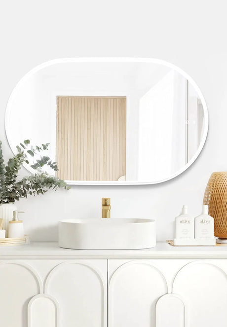 Modern bathroom setup featuring the LED Noosa mirror (900x600) with a white metal frame, mounted above an elegant white vanity with arched panel detailing. The setup includes a minimalist round basin, a gold faucet, and natural accents such as eucalyptus, wicker, and al.ive hand soap bottles.