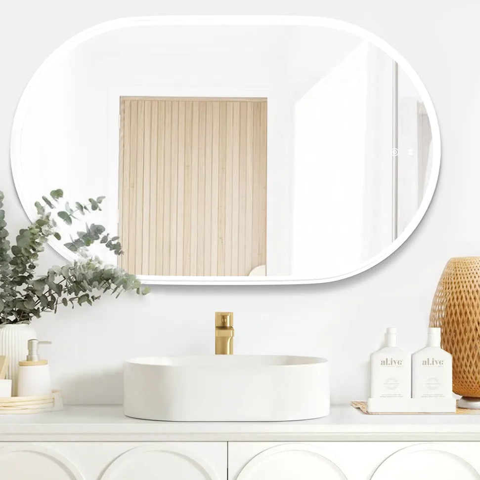 Modern bathroom setup featuring the LED Noosa mirror (900x600) with a white metal frame, mounted above an elegant white vanity with arched panel detailing. The setup includes a minimalist round basin, a gold faucet, and natural accents such as eucalyptus, wicker, and al.ive hand soap bottles.