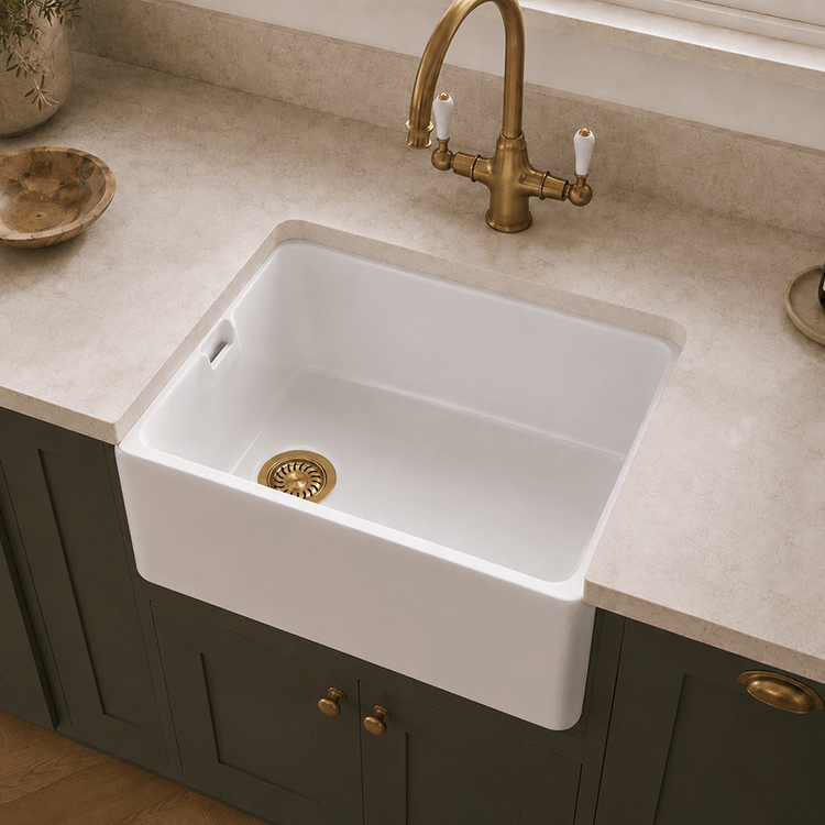A white farmhouse-style undermount sink installed in a beige stone countertop, paired with a brass faucet and dark green cabinets.