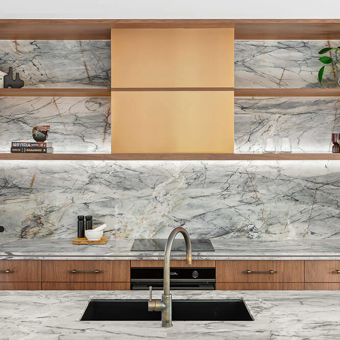 A modern kitchen interior featuring a large marble-patterned backsplash and matching marble countertop, with warm wood cabinetry and open shelving displaying decorative objects.