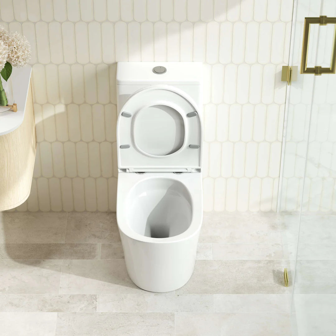 A modern white wall-mounted toilet with the seat lid open, set against a beige hexagonal tile wall and light stone floor. A glass shower door with gold hardware is visible on the right, and part of a vanity with flowers appears on the left.