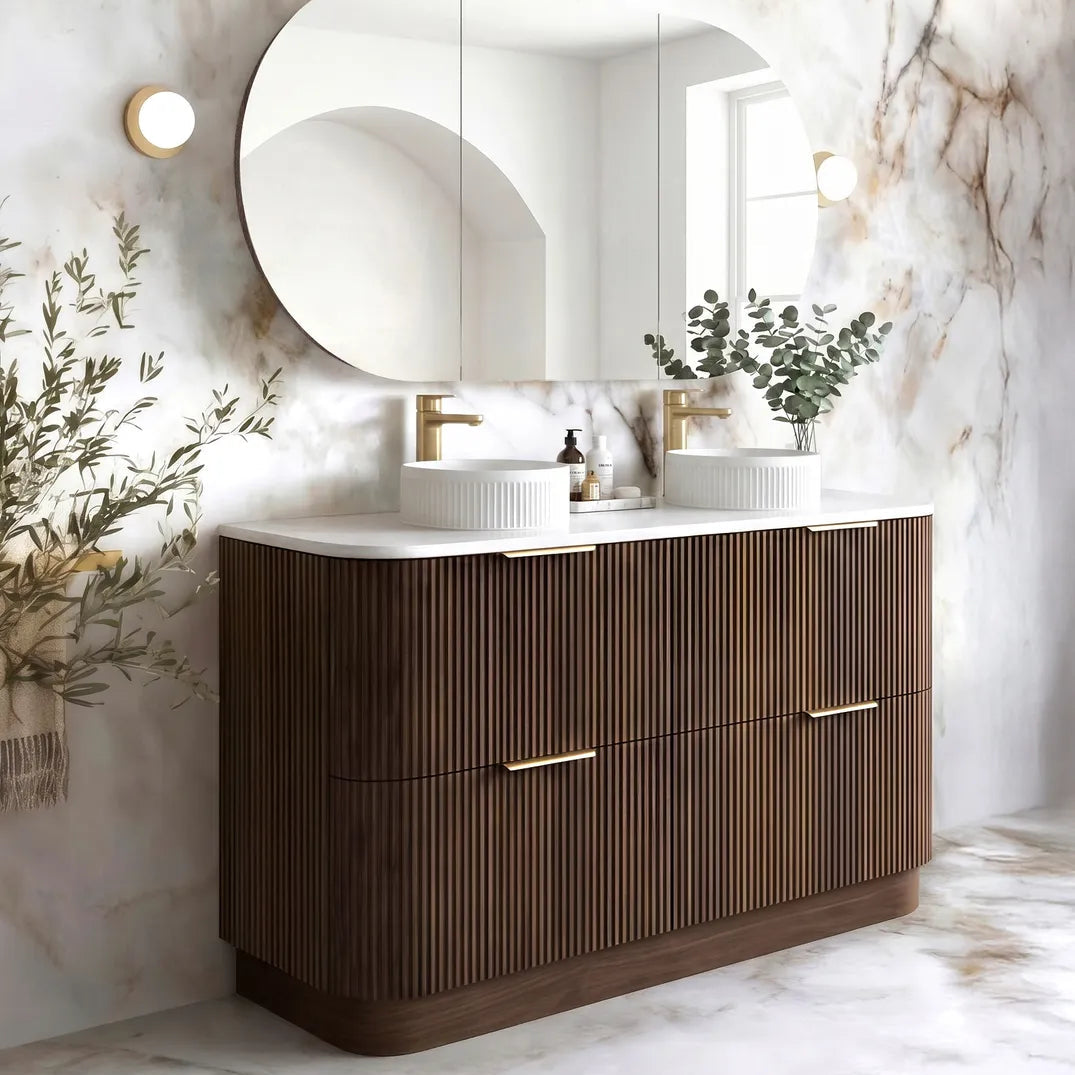 A modern double-sink bathroom vanity with a fluted dark brown wood cabinet, white countertop, and a large round white mirror, set against a marble-patterned wall.
