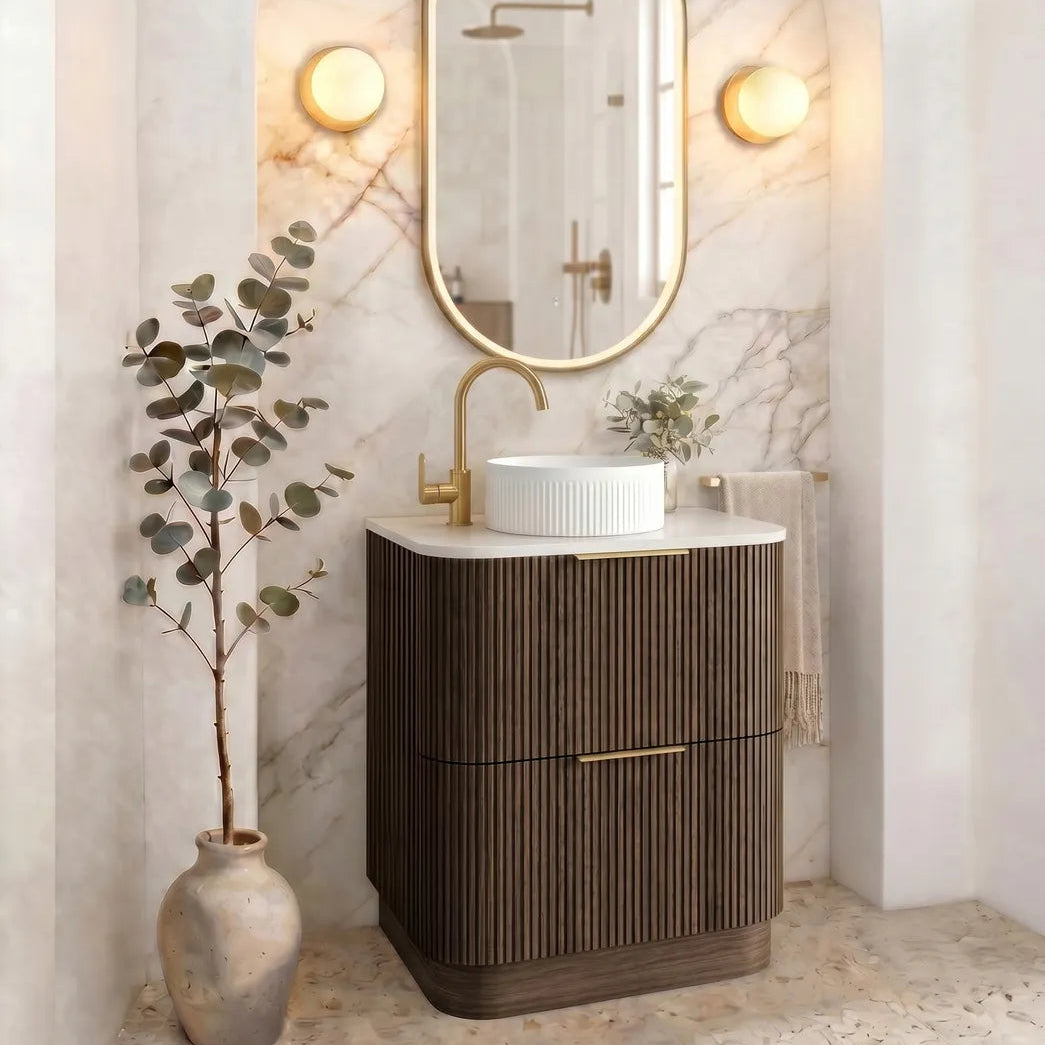 A modern bathroom vanity featuring a dark brown fluted cabinet, a white vessel sink, a gold faucet, and an oval gold-framed mirror set against a marble-look arched wall with warm wall sconces.
