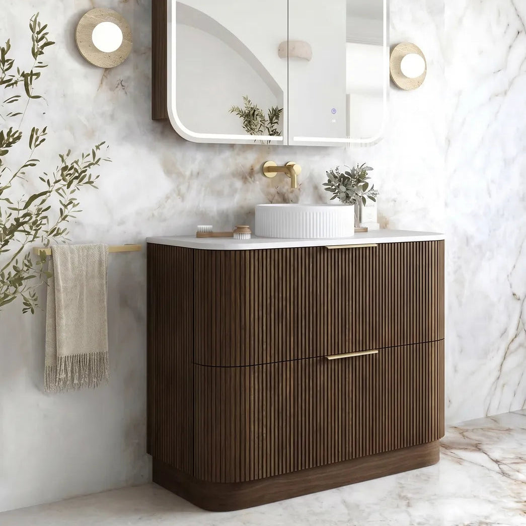 A modern bathroom vanity set featuring a dark brown ribbed cabinet with a white countertop, an integrated white vessel sink, a gold faucet, a wall-mounted double-door medicine cabinet with a lighted frame, and a marble-look wall and floor.