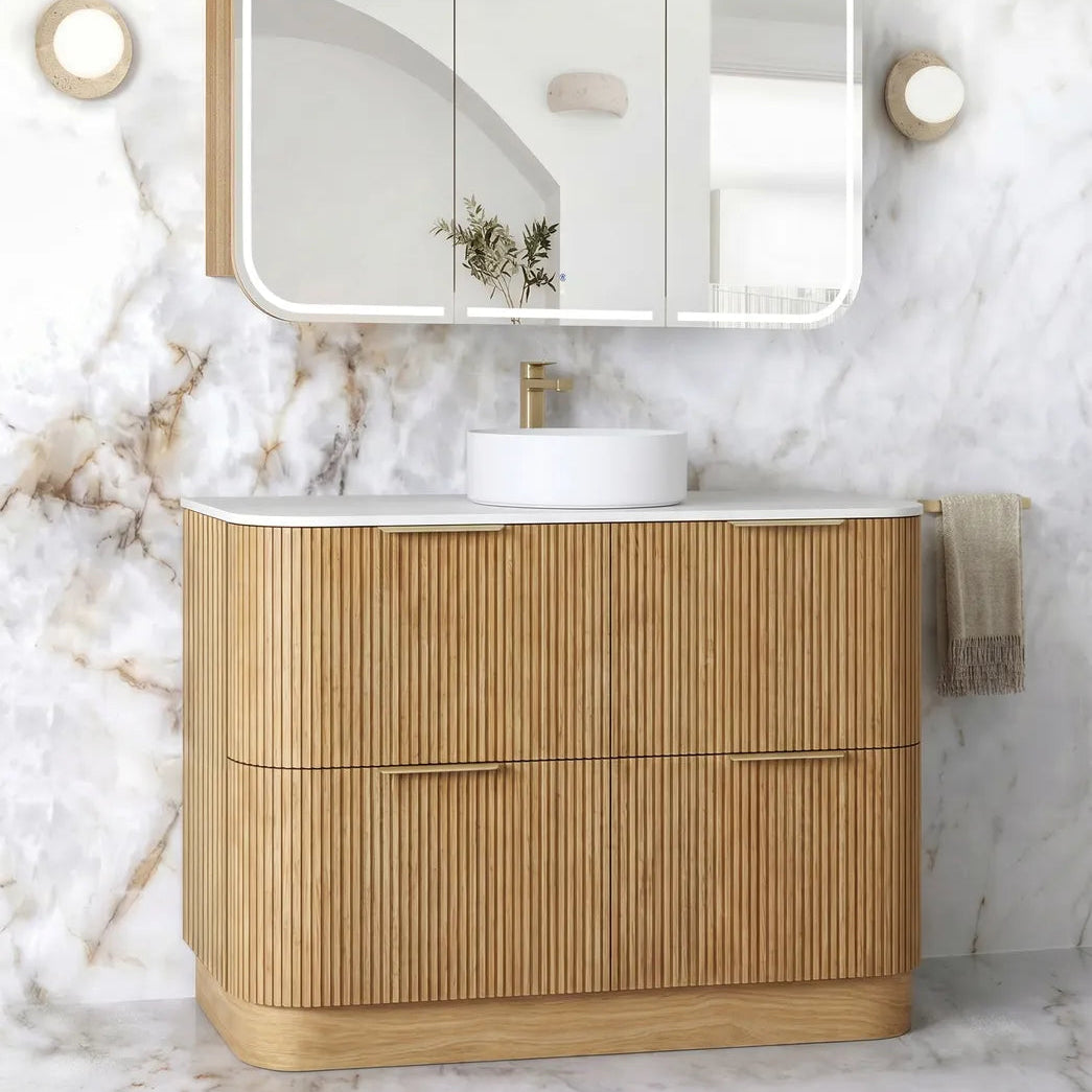 A modern bathroom vanity set featuring a light natural wood ribbed cabinet, a white countertop with a white vessel sink and gold faucet, a three-panel wall-mounted mirror with integrated lighting, and a white marble wall and floor backdrop.