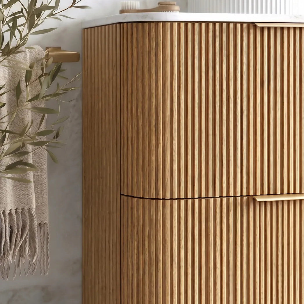 A modern bathroom vanity cabinet with a fluted vertical wood grain finish, featuring a white countertop and brass hardware, shown in a styled bathroom setting with a towel and plant.