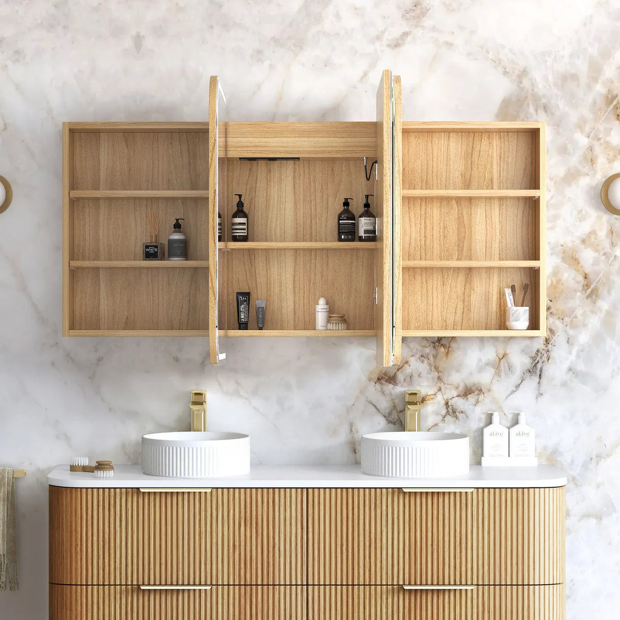 A wall-mounted wooden bathroom mirror cabinet with open doors, styled above a double-sink vanity in a bright bathroom with a marble wall and gold fixtures.