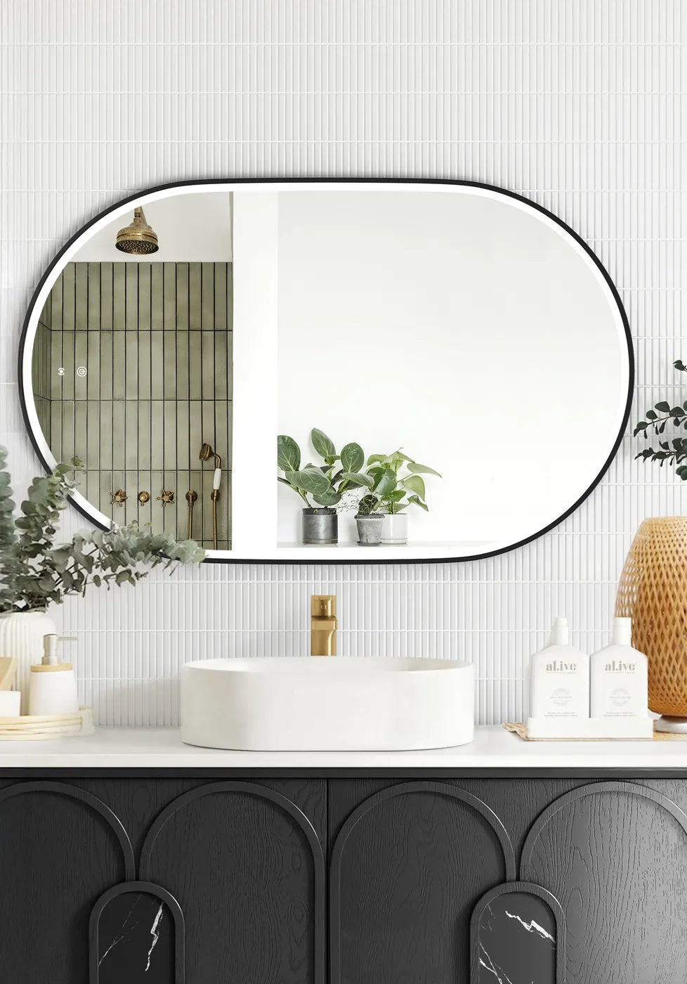 LED Noosa mirror (1200x750) with a sleek black metal frame, mounted above a modern black vanity featuring arched panel detailing. The setup includes a round basin, gold faucet, eucalyptus décor, and al.ive soap bottles, blending modern and minimalist styles.