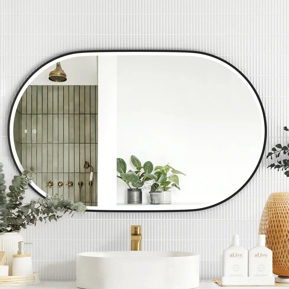 LED Noosa mirror (1200x750) with a sleek black metal frame, mounted above a modern black vanity featuring arched panel detailing. The setup includes a round basin, gold faucet, eucalyptus décor, and al.ive soap bottles, blending modern and minimalist styles.