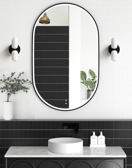 LED Noosa oval mirror (1200x750) with a black metal frame, framed against a textured black tile backsplash. The setup features a marble countertop, round basin, and wall-mounted black fixtures, emphasizing a monochromatic modern aesthetic.