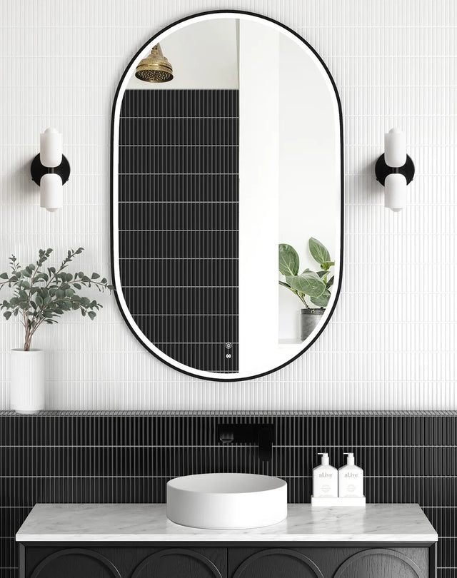 LED Noosa oval mirror (1200x750) with a black metal frame, framed against a textured black tile backsplash. The setup features a marble countertop, round basin, and wall-mounted black fixtures, emphasizing a monochromatic modern aesthetic.