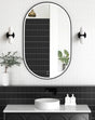 LED Noosa oval mirror (1200x750) with a black metal frame, framed against a textured black tile backsplash. The setup features a marble countertop, round basin, and wall-mounted black fixtures, emphasizing a monochromatic modern aesthetic.