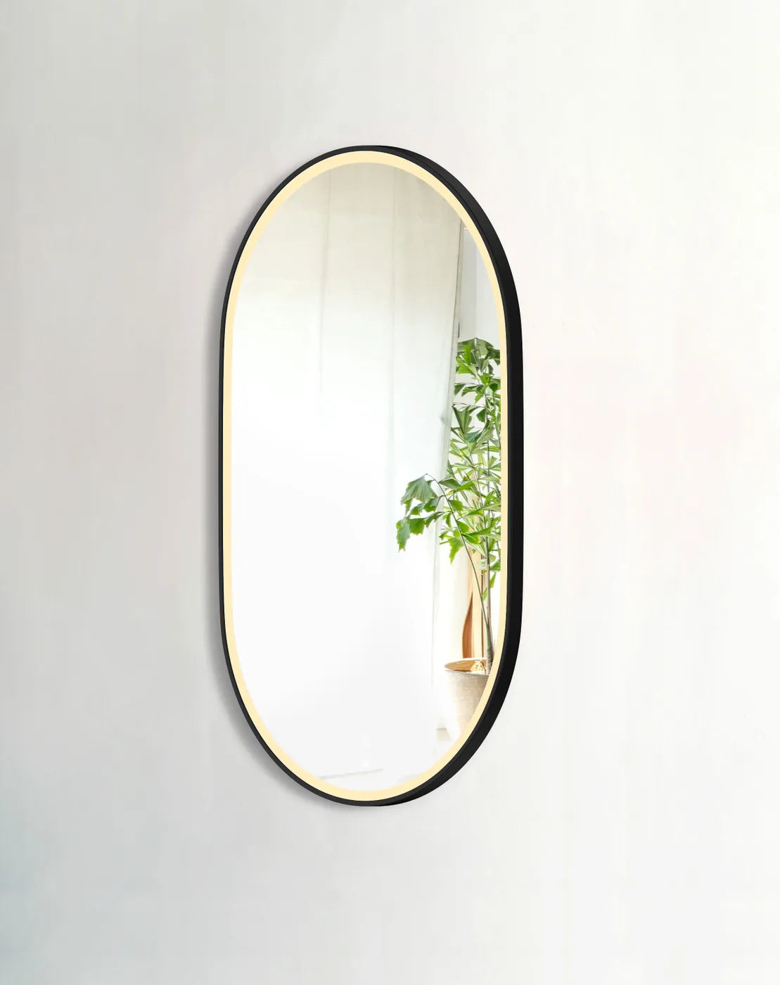 LED Noosa 1200x750 black metal frame mirror, showcasing soft warm backlighting. The mirror is mounted on a neutral wall, reflecting greenery and complementing minimalist décor for a stylish and contemporary bathroom look.