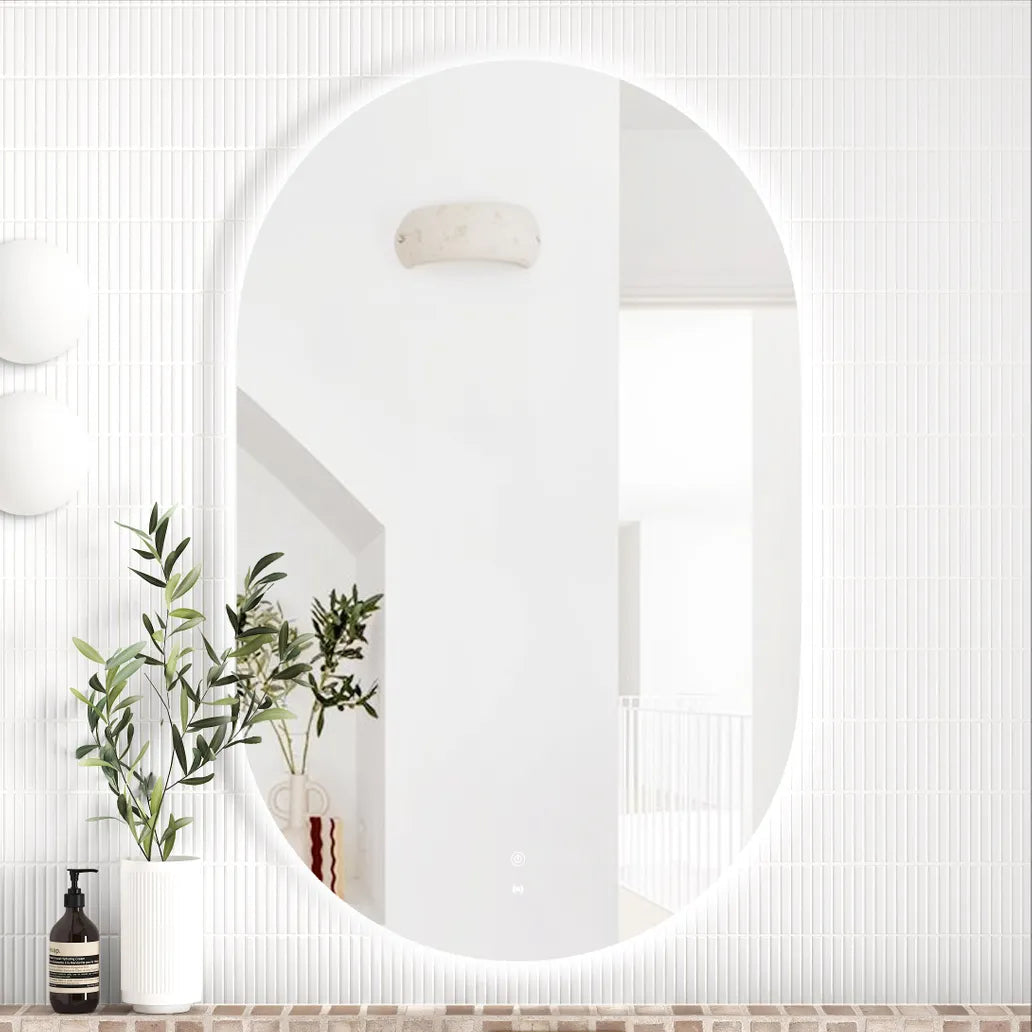 LED Noosa frameless mirror (1200x750) with a clean, curved design, mounted above a vanity with a brick-textured backsplash. The setup highlights a gold faucet, white basin, and a combination of modern and natural decorative elements.