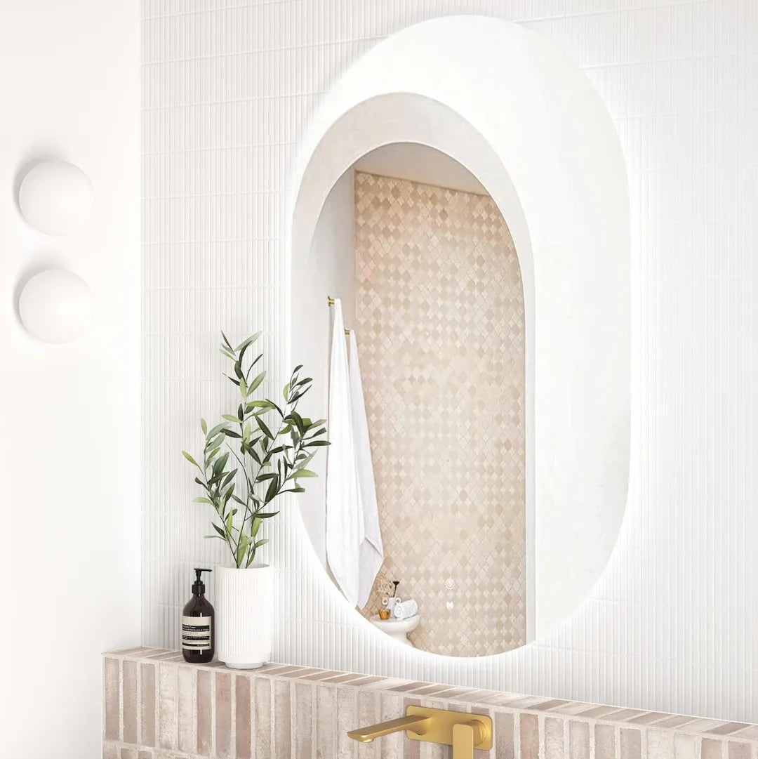 Side view of the LED Noosa 1200x750 frameless mirror, emphasizing its curved edges and ambient lighting. The bathroom includes a white basin, gold faucet, and a brick backsplash, blending warm and contemporary tones.