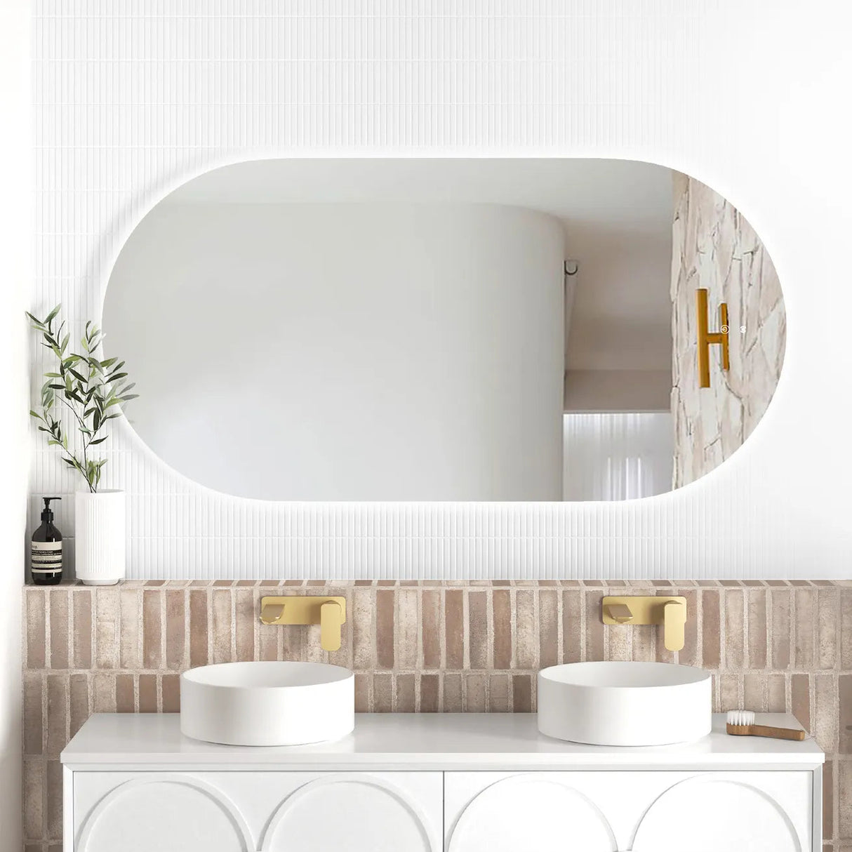 LED Noosa frameless mirror (1500x750) with soft backlighting, mounted above a modern vanity. The bathroom setup features a textured brick backsplash, dual round basins, gold wall-mounted faucets, and minimalist décor including a white vase with greenery.