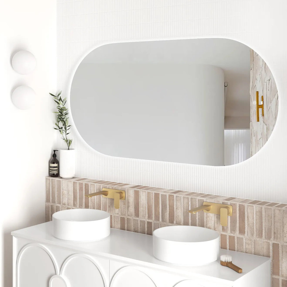 Side angle of the LED Noosa frameless mirror (1500x750), showcasing soft backlighting and a clean, modern bathroom design. The setup includes dual basins, gold wall-mounted faucets, and accent lighting with white globe sconces.