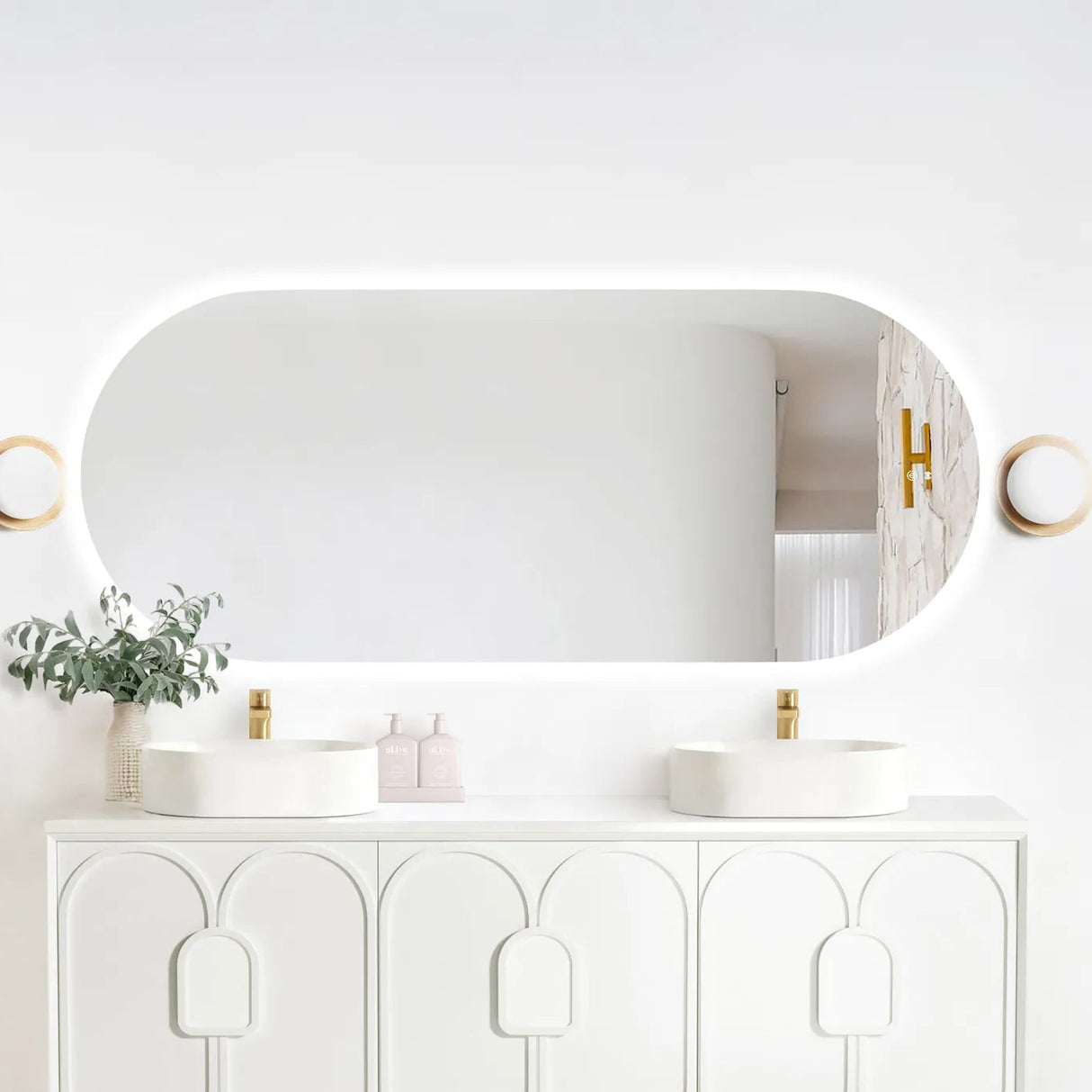 LED Noosa frameless mirror (1800x750) with soft backlighting, mounted above a sleek white vanity featuring arched panel detailing. The setup includes dual round basins, gold faucets, and minimalist accents such as eucalyptus in a textured vase, modern lighting fixtures, and al.ive soap bottles for a clean, contemporary bathroom aesthetic.