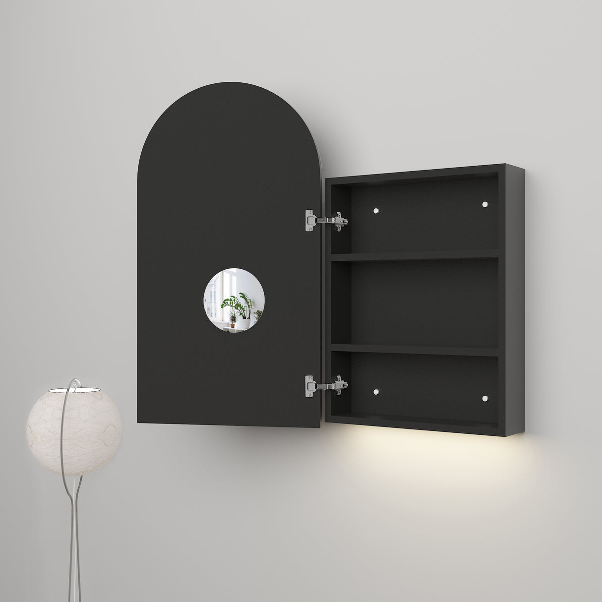 Modern black wall-mounted cabinet with an arched door, featuring a circular cutout that reveals a view of a plant inside. The interior includes two shelves for storage, and the cabinet is paired with soft ambient lighting underneath and a minimalist floor lamp nearby.