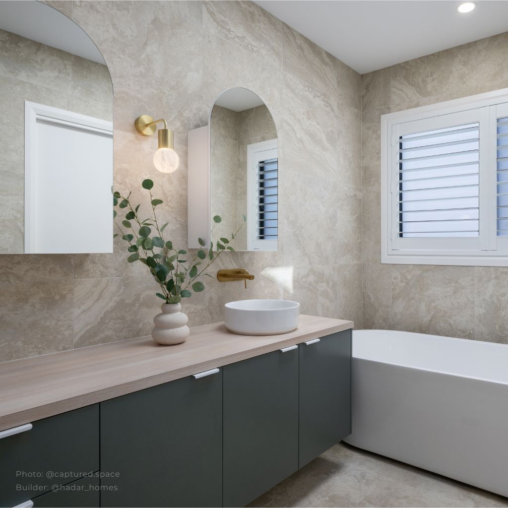 Modern bathroom featuring a wood countertop vanity with dark green cabinets, a round white basin, and a gold wall-mounted faucet. Beige marble-effect tiles, arched mirrors with a gold sconce, and a freestanding white bathtub create a warm and elegant atmosphere, accented by a ceramic vase with eucalyptus branches.