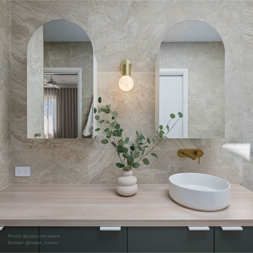 Elegant bathroom design featuring dual arched mirrors with a central gold sconce light mounted on beige marble-effect wall tiles. A light wood vanity top holds a modern white oval countertop basin paired with a gold wall-mounted faucet. A decorative ceramic vase with eucalyptus branches adds a natural and stylish touch.