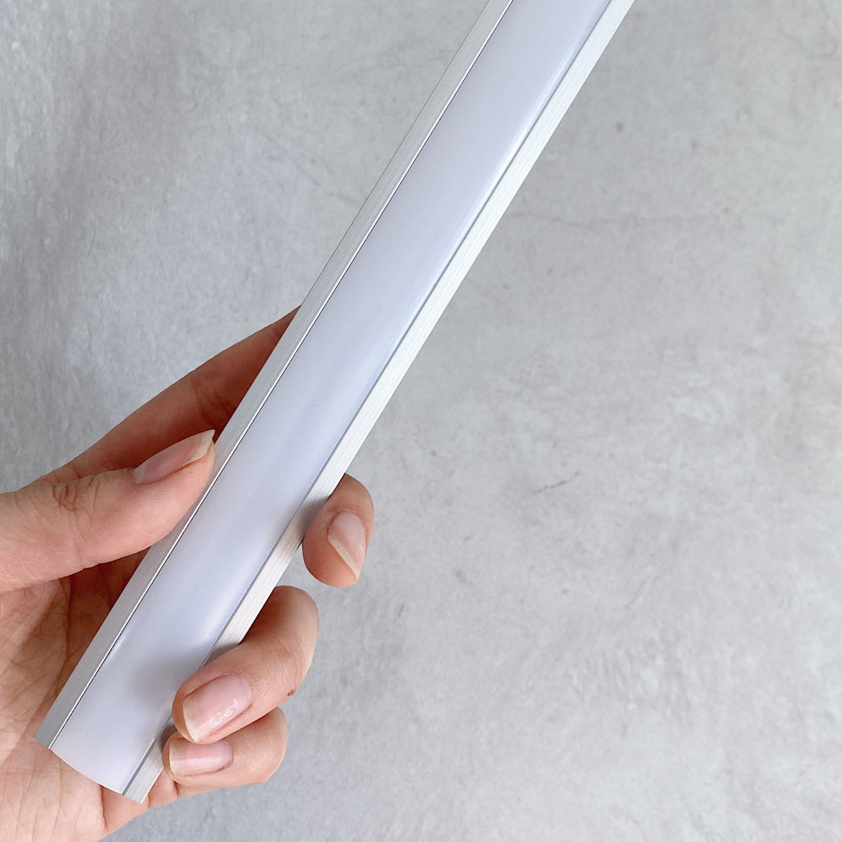 Close-up of a hand holding a slim aluminum light channel with a frosted diffuser cover, showcasing its sleek and modern design against a light gray textured background.