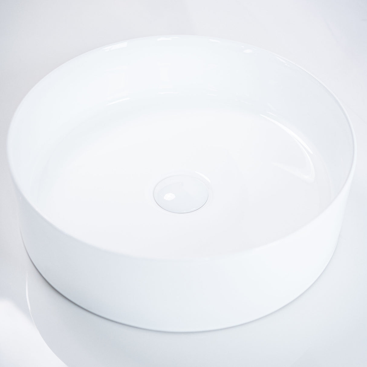 Angled view of a sleek round white ceramic basin with smooth edges and a central drain, set against a clean white background to emphasize its minimalist design.