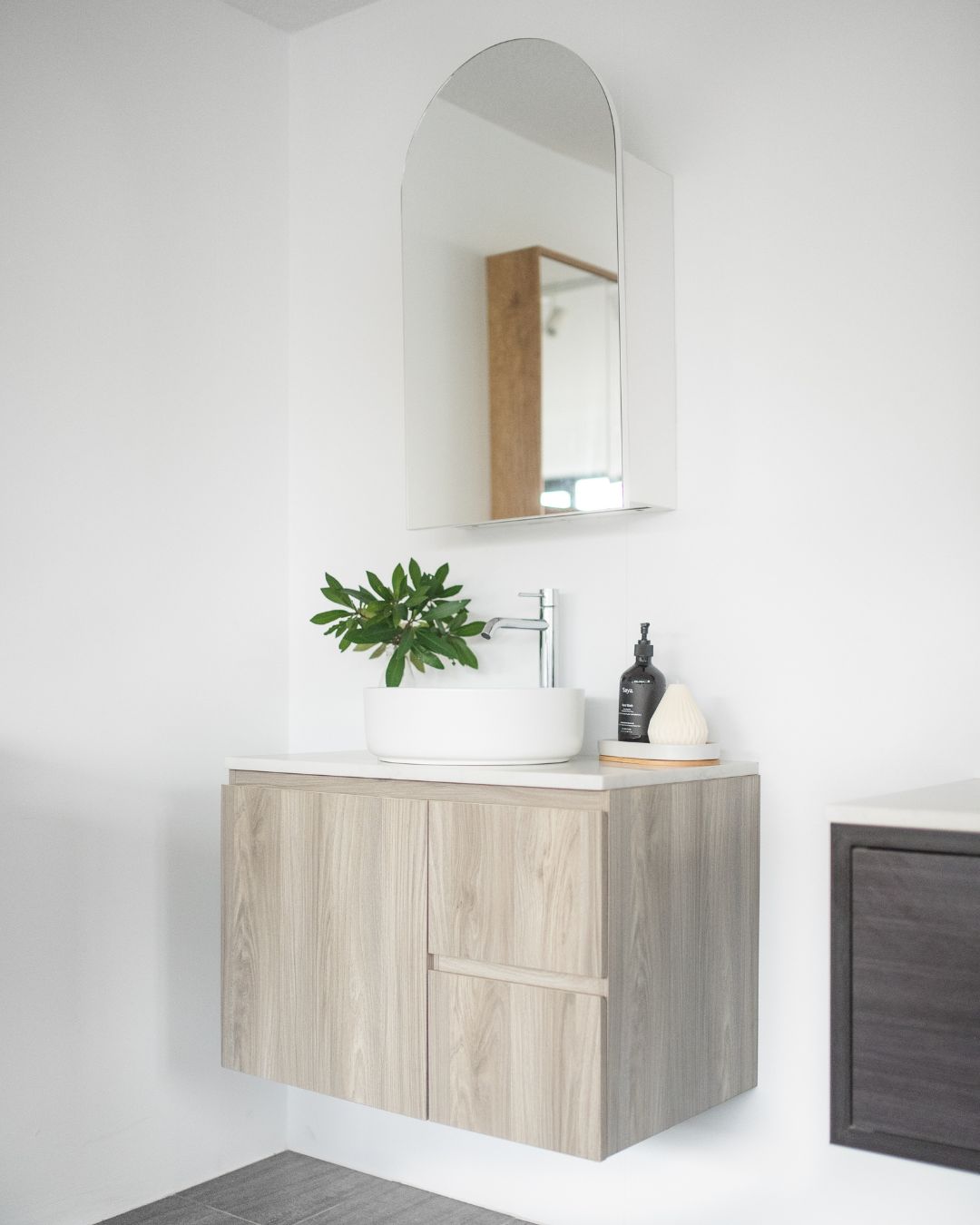 Minimalist bathroom setup featuring a floating light wood vanity with a white round countertop basin and a chrome faucet. An arched mirror hangs above, reflecting a modern aesthetic. The vanity is styled with a green leafy plant, a black soap dispenser, and a small white decorative accessory placed on a wooden tray, all against a clean white background.