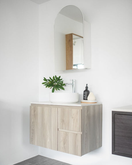 Minimalist bathroom setup featuring a floating light wood vanity with a white round countertop basin and a chrome faucet. An arched mirror hangs above, reflecting a modern aesthetic. The vanity is styled with a green leafy plant, a black soap dispenser, and a small white decorative accessory placed on a wooden tray, all against a clean white background.