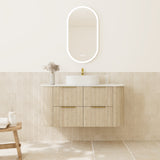 A modern bathroom vanity with a light wood floating cabinet, white vessel sink, gold faucet, and an oval backlit mirror on a cream wall above beige tile.