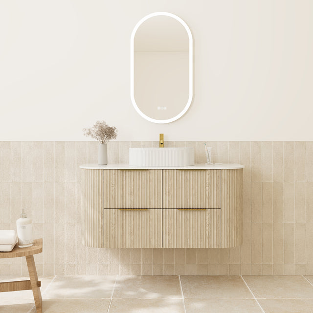 A modern bathroom vanity with a light wood floating cabinet, white vessel sink, gold faucet, and an oval backlit mirror on a cream wall above beige tile.