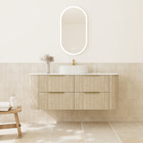 A modern floating bathroom vanity with a ribbed light wood cabinet, white countertop, white vessel sink, and a glowing oval LED mirror mounted on a beige tiled wall.