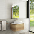 Modern bathroom with wooden vanity, white sink, and large mirror reflecting greenery.