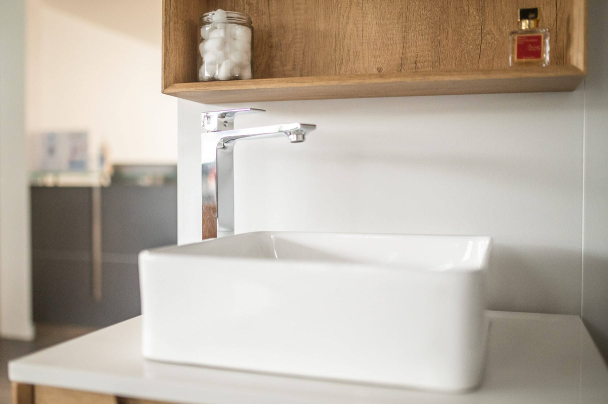 Modern rectangular white ceramic sink with a sleek chrome faucet, set against a wooden shelf holding a jar of cotton balls and a small red perfume bottle.