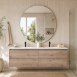 A modern floating double-sink bathroom vanity in light oak wood, paired with a large round wall mirror, black faucets, and a white marble countertop in a bright, tiled bathroom.