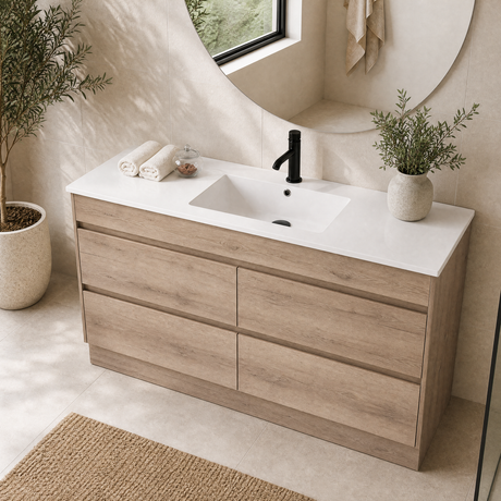 A modern bathroom vanity with a light oak wood cabinet, a white countertop, and an integrated white rectangular sink. The vanity is styled in a bright bathroom with a black faucet, a round mirror, potted plants, and a woven rug.