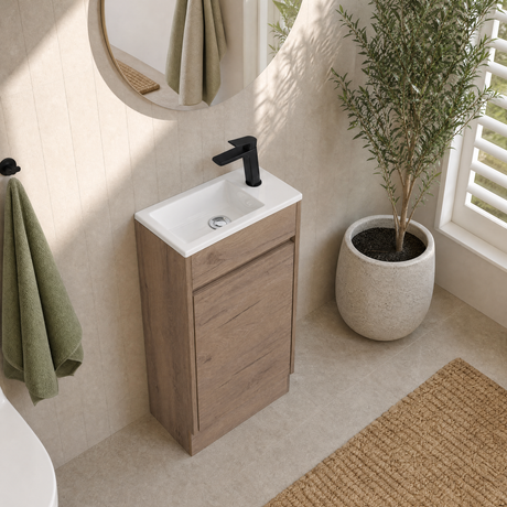A modern single-sink vanity with a light oak wood cabinet and a white ceramic basin, paired with a matte black faucet. The vanity is styled in a bright bathroom setting with a round mirror, a hanging green towel, a potted plant, and a woven rug.