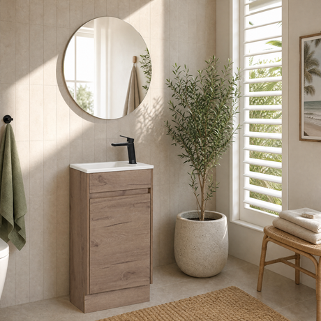 A modern bathroom vanity with a light oak wood cabinet, white sink basin, and matte black faucet, styled in a bright bathroom with a round mirror, potted plant, and window shutters.