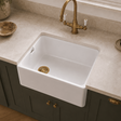 A white farmhouse-style undermount sink installed in a beige stone countertop, paired with a brass faucet and dark green cabinets.