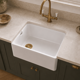 A white farmhouse-style undermount sink installed in a beige stone countertop, paired with a brass faucet and dark green cabinets.