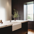 A modern kitchen island with a white farmhouse sink, a matte black gooseneck faucet, and a vase of greenery, set against dark wood cabinetry and a large window.