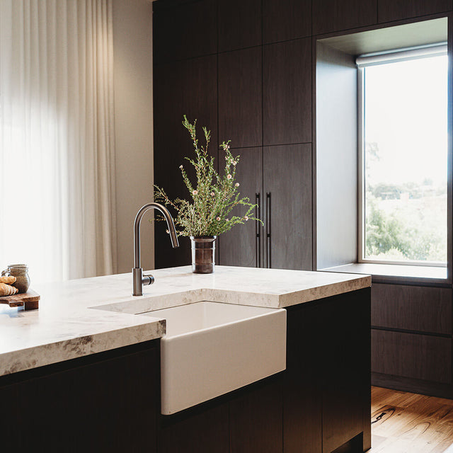 A modern kitchen island with a white farmhouse sink, a matte black gooseneck faucet, and a vase of greenery, set against dark wood cabinetry and a large window.