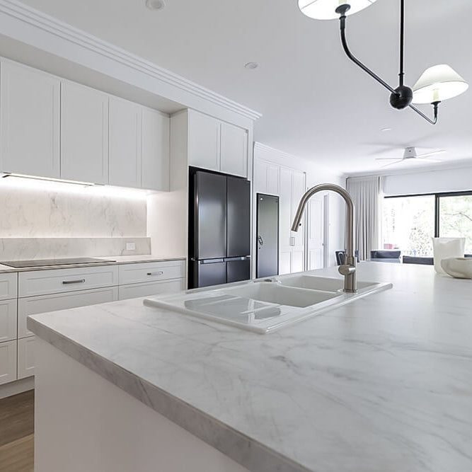 A modern kitchen interior with white cabinetry, a white marble island countertop, a double sink, a curved metallic faucet, and a black refrigerator in the background.