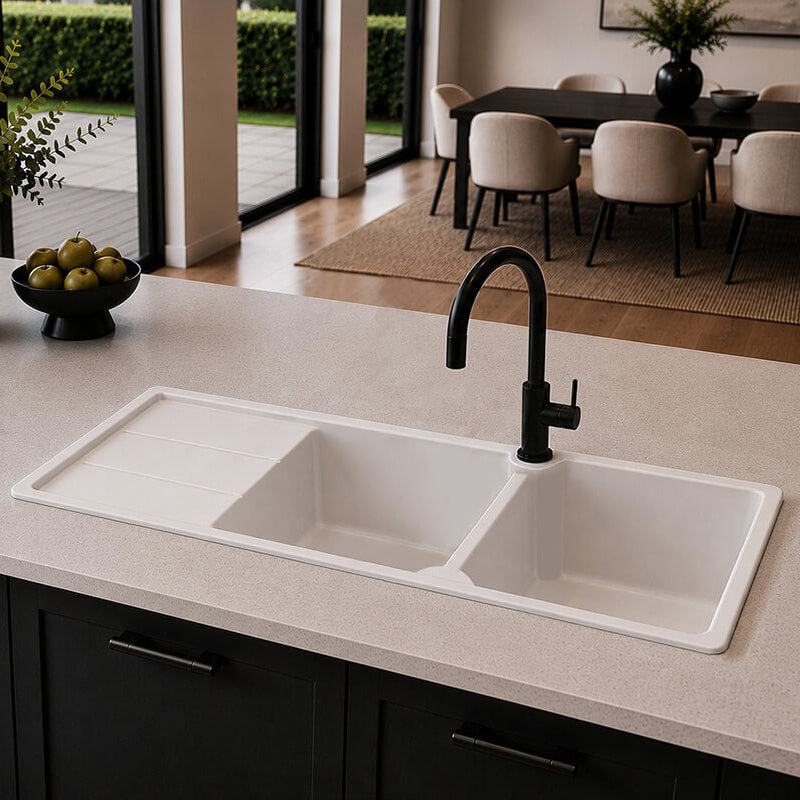 A modern double-bowl white sink with a black gooseneck faucet installed in a light stone countertop, set in a contemporary kitchen with dark cabinets and a dining area in the background.