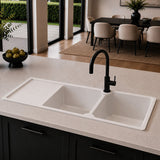 A modern double-bowl white sink with a black gooseneck faucet installed in a light stone countertop, set in a contemporary kitchen with dark cabinets and a dining area in the background.