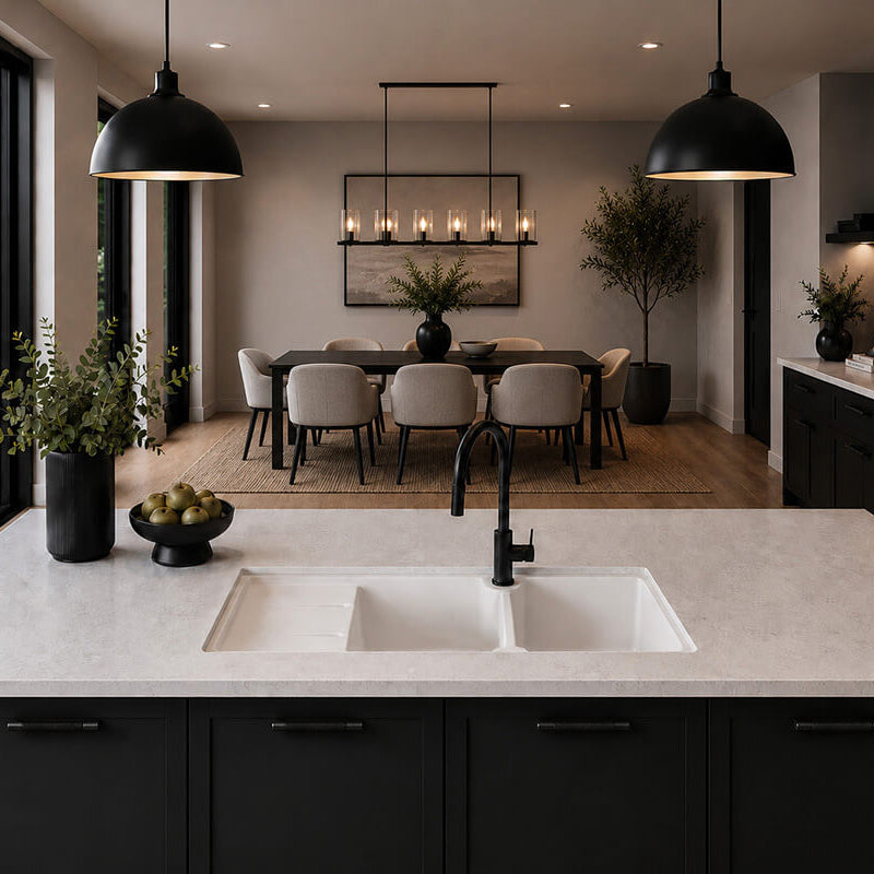 A modern kitchen island with a white double-bowl sink and matte black faucet, set against a dark charcoal cabinetry base. The background reveals a spacious, contemporary dining room with a dark wood table, gray upholstered chairs, and black pendant lights.