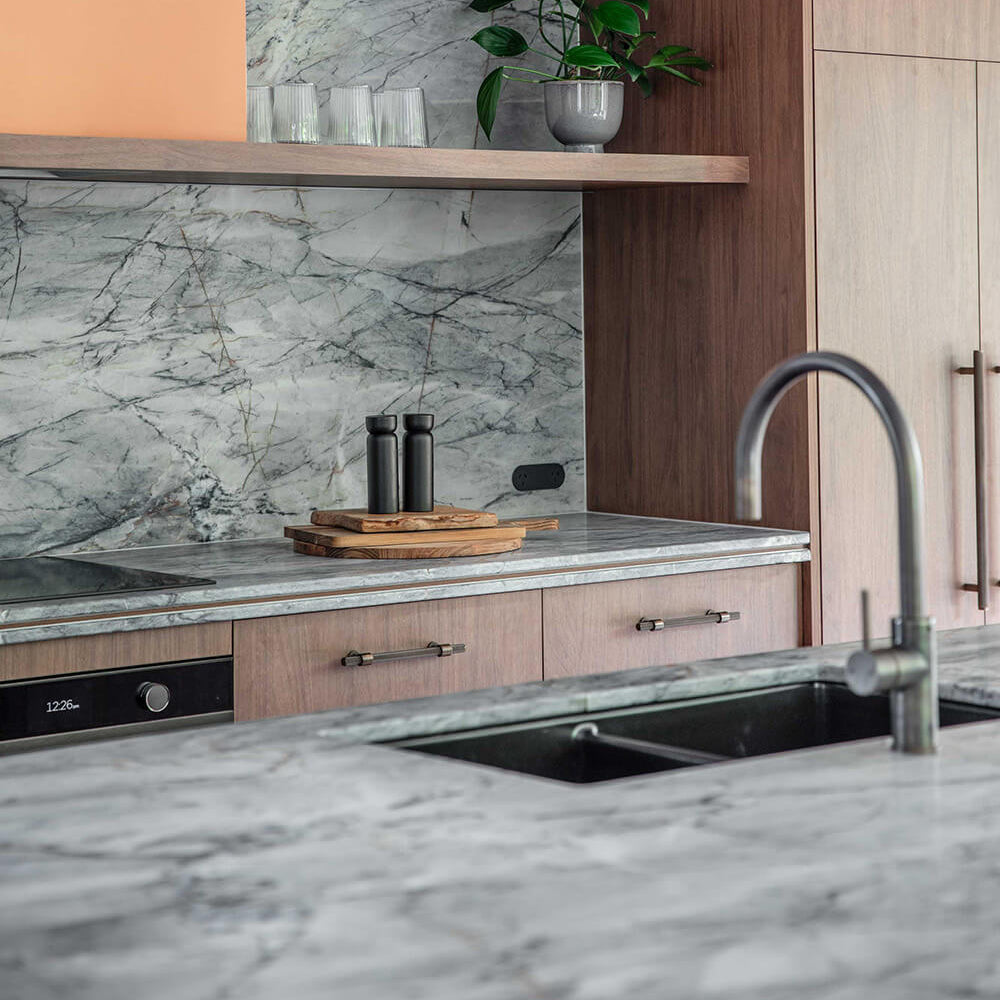 Modern kitchen cabinetry with warm wood panels, a gray marble backsplash and countertop, a black sink, and a curved metallic faucet. An open shelf holds a small green plant, and the scene is photographed in a real interior setting.